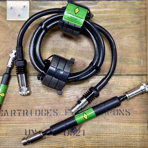 Op de afbeelding: Een zwarte coaxkabel opgerold met twee zwarte ferrietkernen en twee rechte kabelsegmenten. Elk segment is voorzien van zilverkleurige metalen connectoren aan beide uiteinden en een groen label met een geel ruitvormig logo en zwarte tekst.