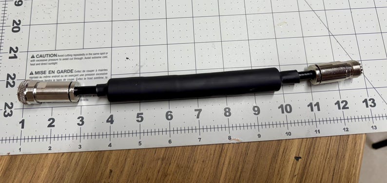 May include: A black cable with silver connectors on each end. The cable is about 10 inches long and is measured on a ruler.