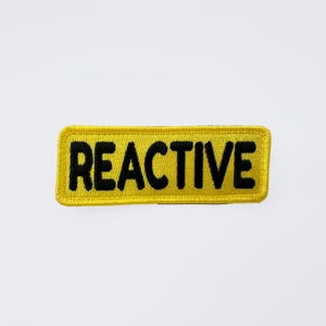 May include: A yellow rectangular patch with the word "REACTIVE" embroidered in black block letters. The patch has a yellow border and is set against a white background. This patch is suitable for jackets, bags, or other fabric items.