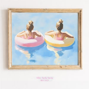May include: A framed print depicts two figures in pink swimsuits, each floating in a pool on a pink and yellow inflatable ring. The artwork is set against a blue water background, with a light-colored wooden frame. The print is from the Cotton Candy Canvas Print Shop.