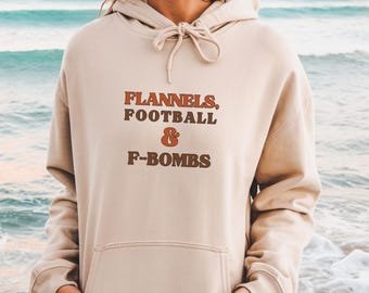 Op de afbeelding: Beige hoodie met een trekkoord en een voorzak. De tekst "FLANNELS, FOOTBALL & F-BOMBS" is gedrukt in bruine en oranje letters. De hoodie is gemaakt van een zacht materiaal.