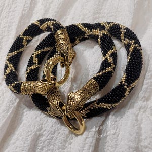 Puede incluir: Dos pulseras circulares hechas de cuentas negras con un patr&oacute;n de diamantes de cuentas doradas. Cada pulsera presenta extremos de metal dorado ornamentados con forma de cabezas de animales, unidos por un cierre de anillo circular pulido.