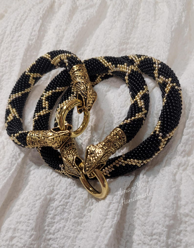 Puede incluir: Dos pulseras circulares hechas de cuentas negras con un patr&oacute;n de diamantes de cuentas doradas. Cada pulsera presenta extremos de metal dorado ornamentados con forma de cabezas de animales, unidos por un cierre de anillo circular pulido.