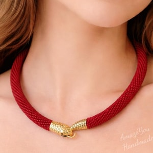 May include: A red beaded necklace with gold-colored snake head clasps. The necklace is a choker style, with a thick, textured appearance. The clasp features a detailed scale pattern. The text "Amaze You Handmade" is visible in the image.