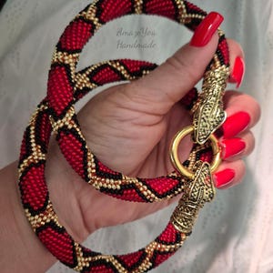 May include: A beaded necklace featuring a red, black, and gold geometric pattern. The necklace has a gold-toned metal clasp shaped like two snake heads connected by a circular ring. The text AmazeYou Handmade is visible on the beaded surface.