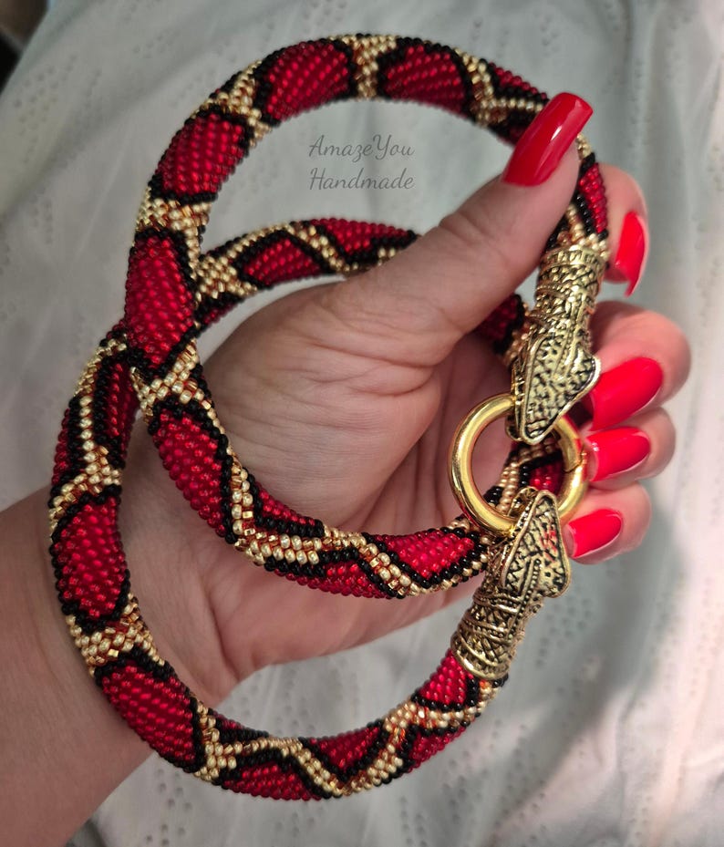 May include: A beaded necklace featuring a red, black, and gold geometric pattern. The necklace has a gold-toned metal clasp shaped like two snake heads connected by a circular ring. The text AmazeYou Handmade is visible on the beaded surface.