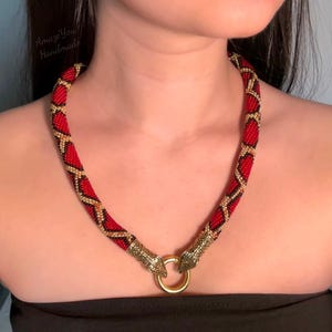 May include: A beaded necklace featuring a red, black, and gold geometric pattern. The necklace ends in two gold-toned metal snake head terminals that connect to a central gold-toned circular ring clasp. The text AmazeYou Handmade is visible.