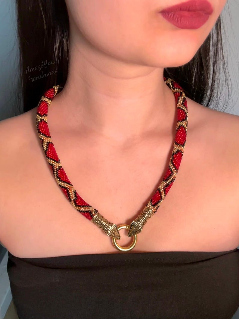 May include: A beaded necklace featuring a red, black, and gold geometric pattern. The necklace ends in two gold-toned metal snake head terminals that connect to a central gold-toned circular ring clasp. The text AmazeYou Handmade is visible.