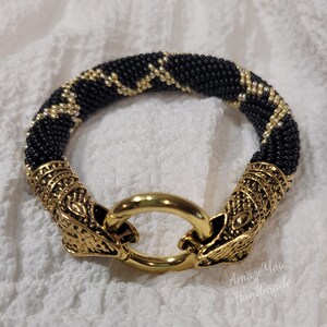 Puede incluir: Una pulsera de cuentas con un patr&oacute;n en zigzag negro y dorado. La pulsera termina en dos adornos de metal dorado con forma de cabeza de animal que se conectan a un cierre de anillo central dorado. El texto Amaze You Handmade es visible.