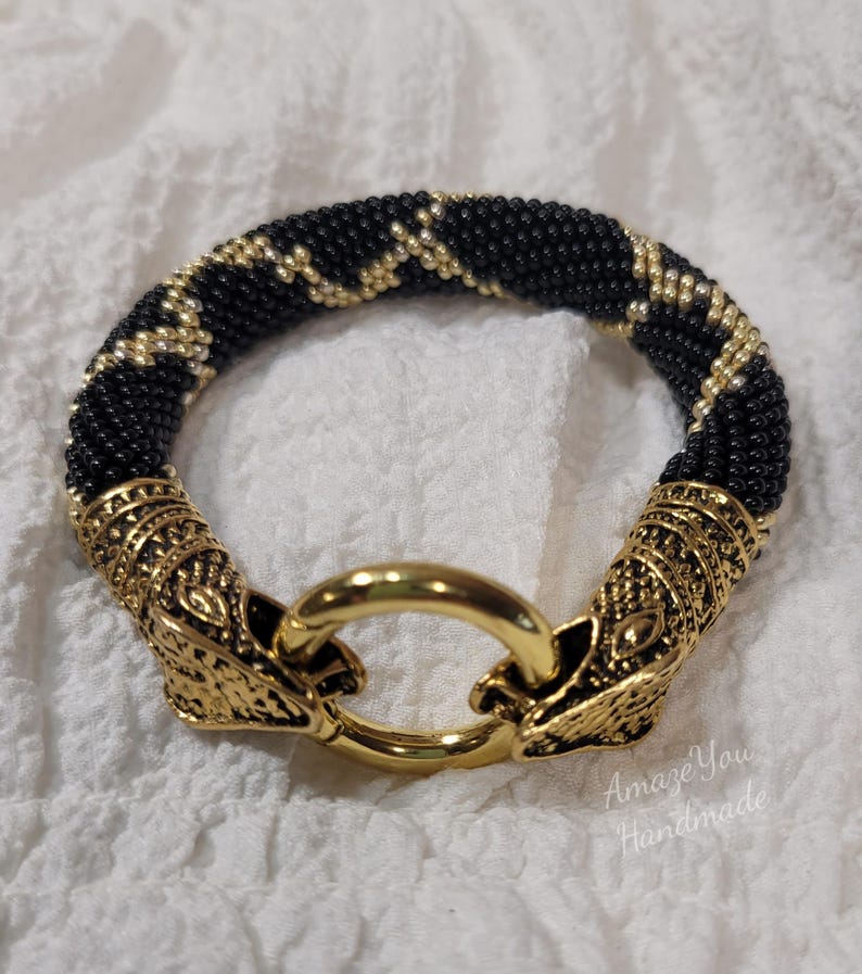 Puede incluir: Una pulsera de cuentas con un patr&oacute;n en zigzag negro y dorado. La pulsera termina en dos adornos de metal dorado con forma de cabeza de animal que se conectan a un cierre de anillo central dorado. El texto Amaze You Handmade es visible.