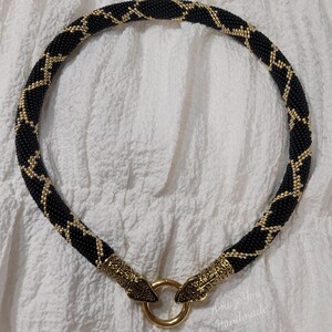 Puede incluir: Un collar de cuentas con un patr&oacute;n geom&eacute;trico en negro y dorado. El collar tiene forma tubular y est&aacute; rematado con dos terminales de metal dorado en forma de cabeza de serpiente que se unen a un cierre de anillo central. El texto AmazeYou Handmade es visible.