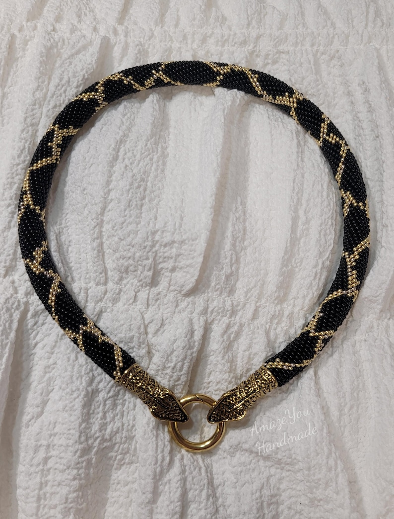 Puede incluir: Un collar de cuentas con un patr&oacute;n geom&eacute;trico en negro y dorado. El collar tiene forma tubular y est&aacute; rematado con dos terminales de metal dorado en forma de cabeza de serpiente que se unen a un cierre de anillo central. El texto AmazeYou Handmade es visible.