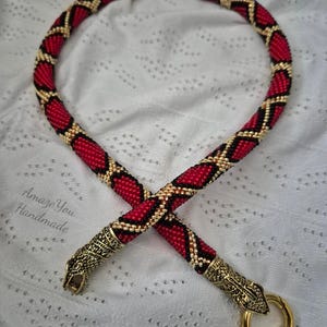 May include: A beaded necklace featuring a red, black, and gold diamond pattern. The ends are finished with metallic gold-toned snake head caps, one of which includes a circular clasp. The text Amaze You Handmade is visible on the left side.