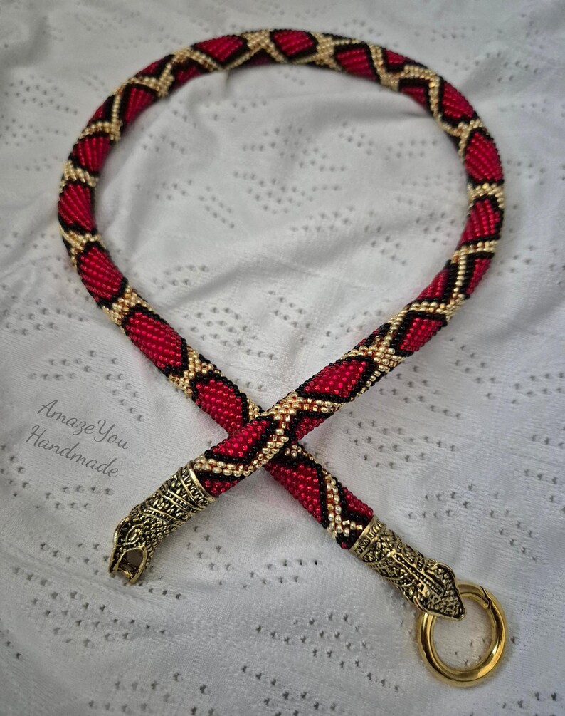 May include: A beaded necklace featuring a red, black, and gold diamond pattern. The ends are finished with metallic gold-toned snake head caps, one of which includes a circular clasp. The text Amaze You Handmade is visible on the left side.