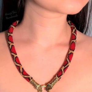 May include: A beaded necklace featuring a red, black, and gold geometric pattern. The necklace ends in two metallic gold-toned snake head clasps that connect to a circular gold-toned ring at the front. The text AmazeYou Handmade is visible.