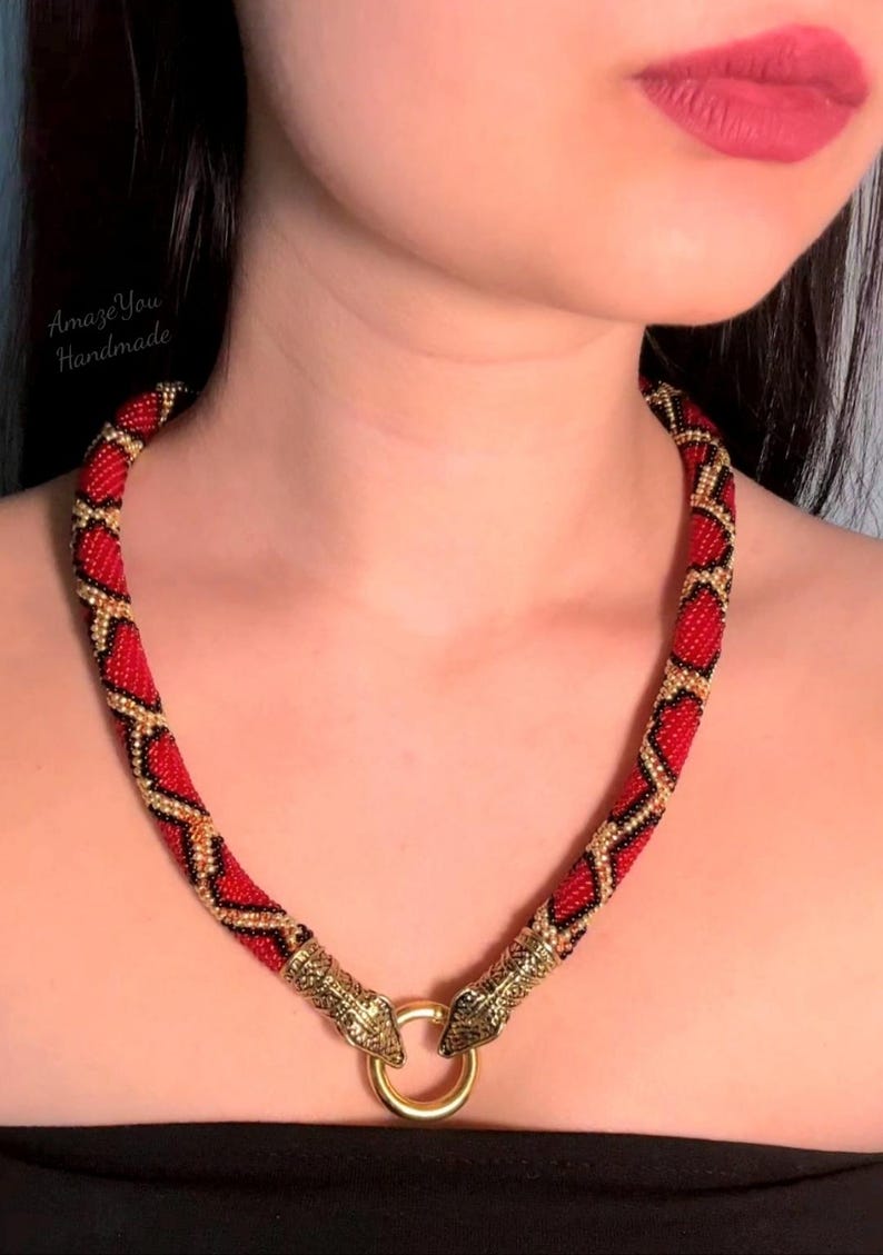 May include: A beaded necklace featuring a red, black, and gold geometric pattern. The necklace ends in two metallic gold-toned snake head clasps that connect to a circular gold-toned ring at the front. The text AmazeYou Handmade is visible.