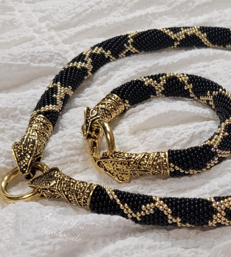 Puede incluir: Un conjunto de collar y pulsera de cuentas con un patr&oacute;n geom&eacute;trico negro y dorado. Las piezas de joyer&iacute;a est&aacute;n terminadas con cierres de metal dorado en forma de cabeza de drag&oacute;n. El texto "Amaze You Handmade" es visible abajo a la izquierda.