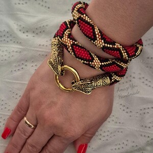 May include: A wrap-around bracelet featuring a beaded snake-like pattern in red, black, and gold. The bracelet is secured with a gold-toned metal clasp shaped like two snake heads meeting at a circular ring. The text Amaze You Handmade is visible.