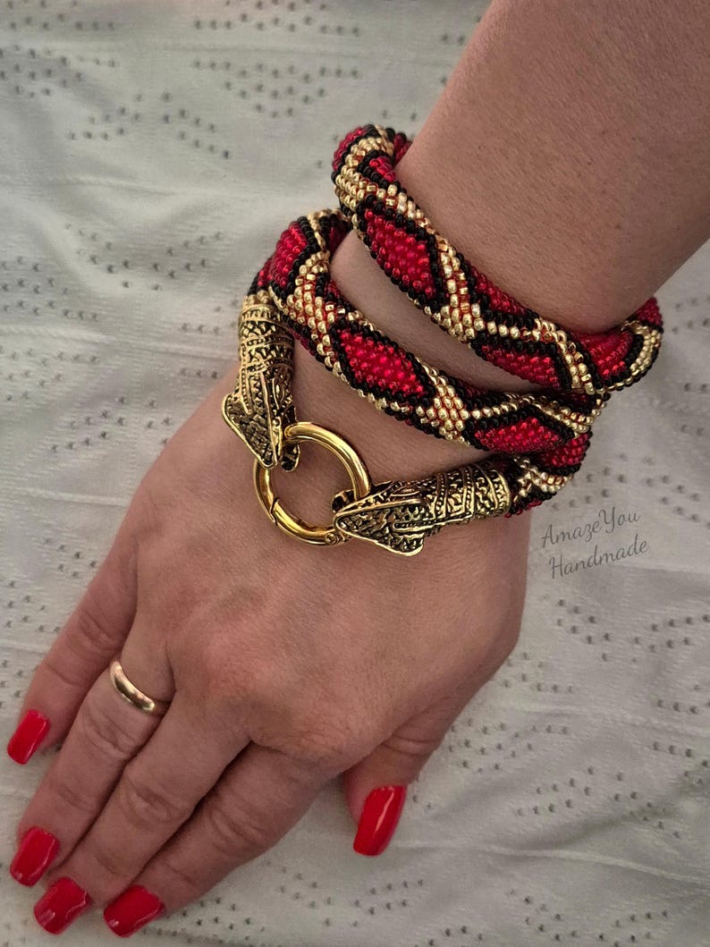 May include: A wrap-around bracelet featuring a beaded snake-like pattern in red, black, and gold. The bracelet is secured with a gold-toned metal clasp shaped like two snake heads meeting at a circular ring. The text Amaze You Handmade is visible.
