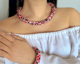 May include: A beaded necklace and matching bracelet set featuring a geometric pattern in red, black, and white seed beads. The cylindrical jewelry pieces display a repeating diamond motif. The text AmazeYou Handmade is visible in the corner.