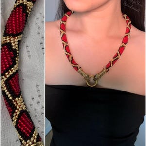 May include: A beaded necklace featuring a repeating diamond pattern in red, black, and gold seed beads. The necklace ends in two metallic gold-toned snake head terminals that connect to a central gold-toned ring clasp. The text Amaze You Handmade is visible.