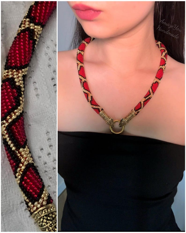 May include: A beaded necklace featuring a repeating diamond pattern in red, black, and gold seed beads. The necklace ends in two metallic gold-toned snake head terminals that connect to a central gold-toned ring clasp. The text Amaze You Handmade is visible.
