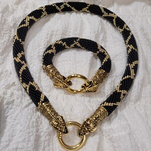 Puede incluir: Un conjunto de collar y pulsera de cuentas con un patr&oacute;n de zigzag en negro y dorado. Ambas piezas est&aacute;n hechas de peque&ntilde;as cuentas negras con detalles dorados y terminadas con cierres de metal dorado en forma de cabeza de serpiente y anillos.