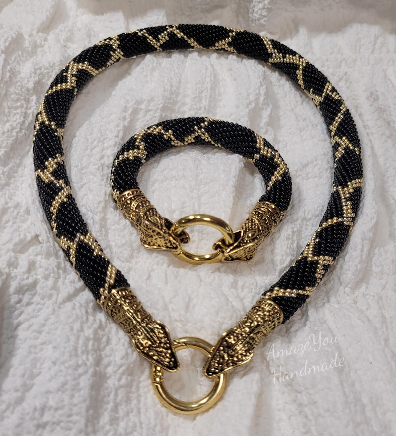 Puede incluir: Un conjunto de collar y pulsera de cuentas con un patr&oacute;n de zigzag en negro y dorado. Ambas piezas est&aacute;n hechas de peque&ntilde;as cuentas negras con detalles dorados y terminadas con cierres de metal dorado en forma de cabeza de serpiente y anillos.