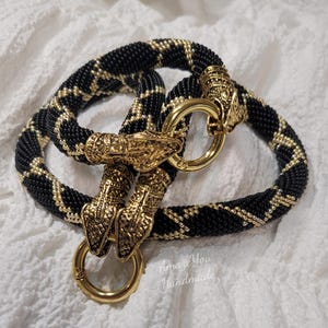 Puede incluir: Un collar de cuentas con un patr&oacute;n geom&eacute;trico negro y dorado, acabado con dos cierres de metal dorado en forma de cabeza de serpiente y dos anillos circulares dorados. El texto Amaze You Handmade es visible en la esquina inferior derecha.