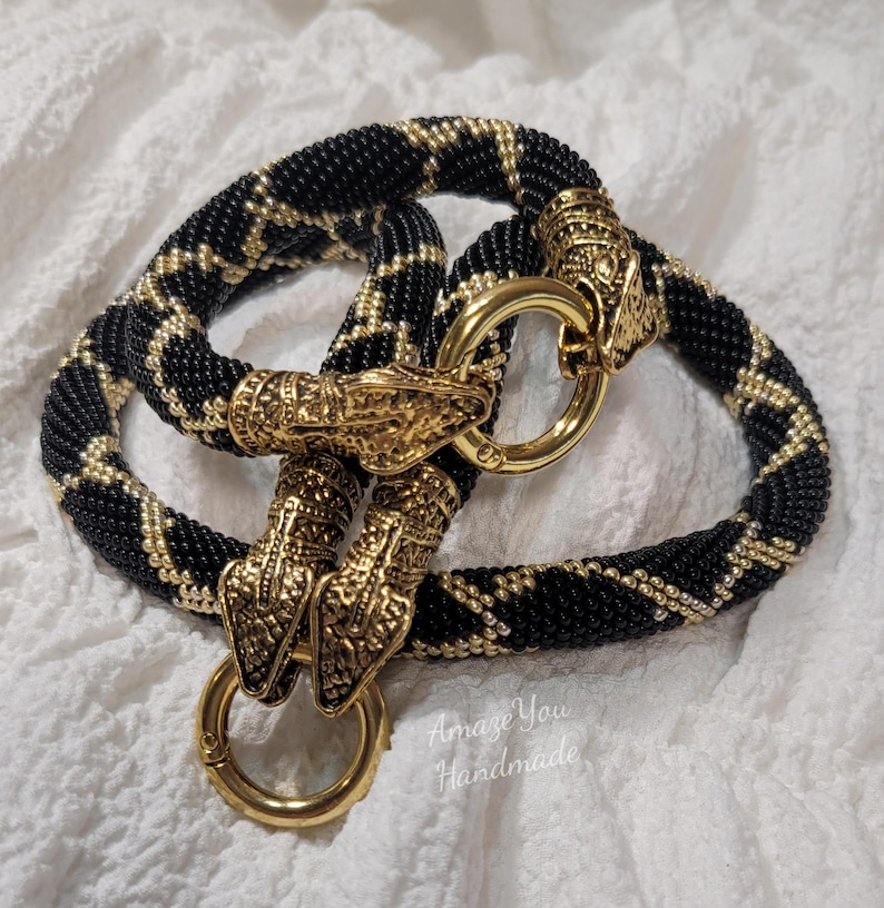 Puede incluir: Un collar de cuentas con un patr&oacute;n geom&eacute;trico negro y dorado, acabado con dos cierres de metal dorado en forma de cabeza de serpiente y dos anillos circulares dorados. El texto Amaze You Handmade es visible en la esquina inferior derecha.
