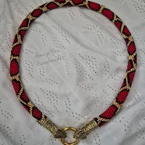 May include: A beaded necklace featuring a red and black diamond pattern with gold accents. The ends are finished with ornate gold-toned metal snake heads that clasp onto a circular gold ring. The text AmazeYou Handmade is visible.
