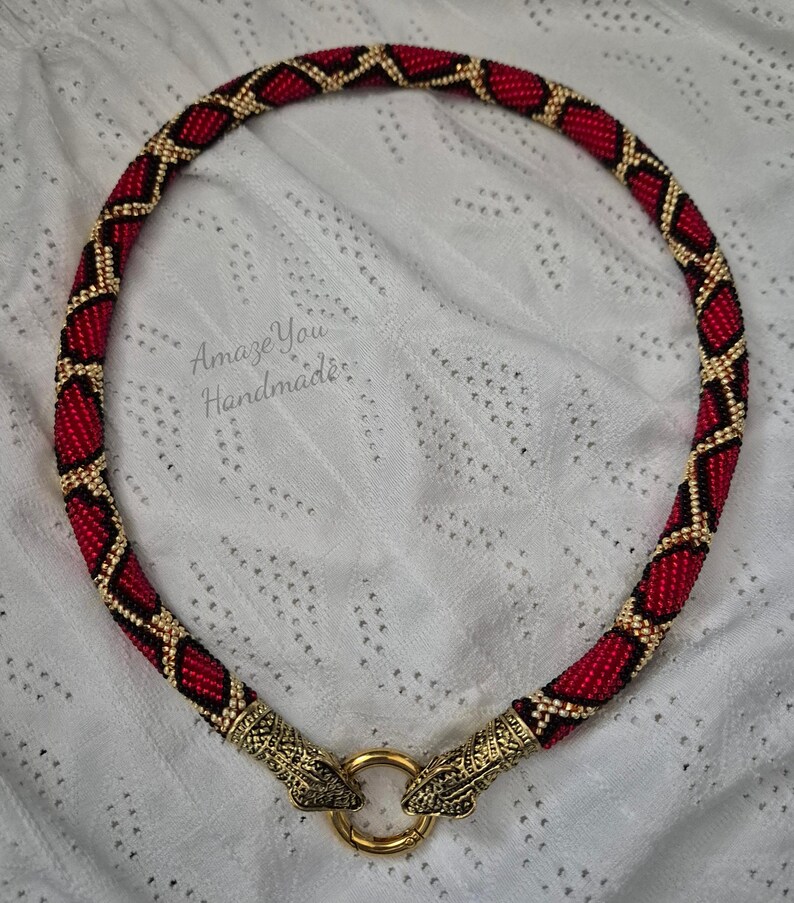 May include: A beaded necklace featuring a red and black diamond pattern with gold accents. The ends are finished with ornate gold-toned metal snake heads that clasp onto a circular gold ring. The text AmazeYou Handmade is visible.
