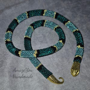 Puede incluir: Un collar de serpiente de cuentas en verde azulado, negro y dorado. El cuerpo de la serpiente es verde azulado con rayas negras y detalles dorados. La cabeza y la cola son de metal dorado. El texto "Amaze You Handmade" está en la imagen.
