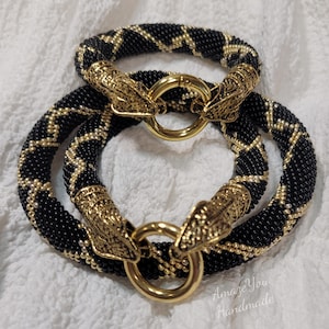 May include: Two circular bracelets crafted from black and gold seed beads in a zigzag pattern. Each bracelet features two ornate gold-toned metal snake head terminals that meet at a central gold-toned circular clasp. Text reads Amaze You Handmade.