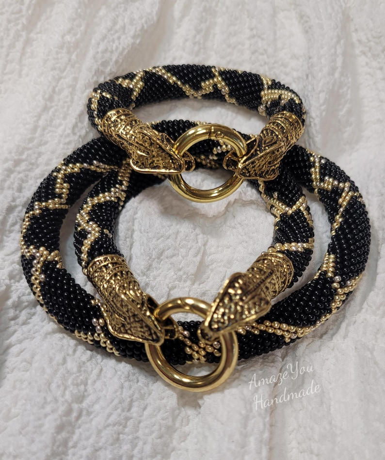 Puede incluir: Dos pulseras circulares hechas de cuentas negras y doradas en un patr&oacute;n de zigzag. Cada pulsera presenta dos terminales de metal dorado con forma de cabeza de serpiente que se unen en un cierre circular central. El texto dice Amaze You Handmade.