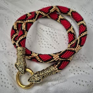 May include: A tubular beaded necklace featuring a repeating diamond pattern in red, black, and gold seed beads. The ends are finished with ornate gold-tone metal snake head caps that connect to a circular gold-tone ring clasp. Text reads Amaze You Handmade.