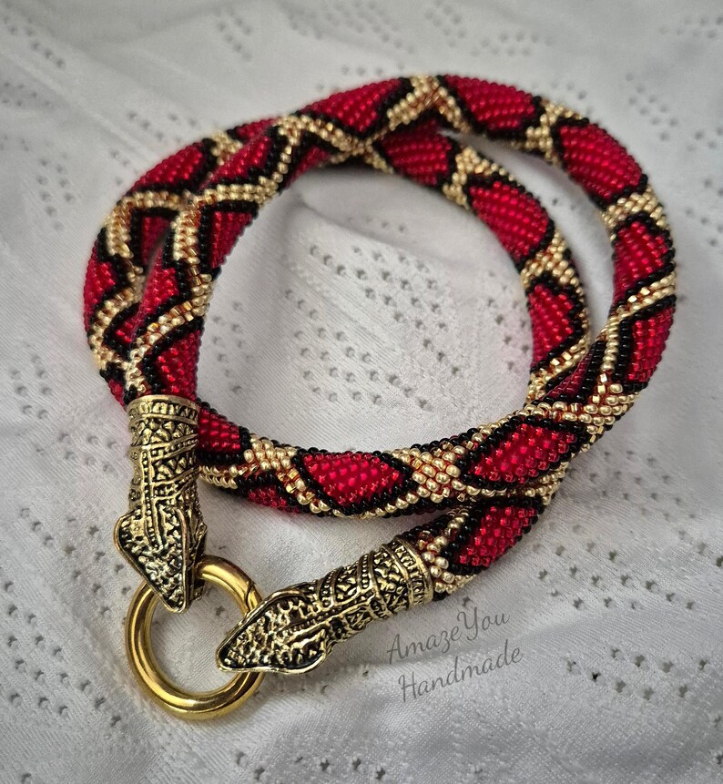 May include: A tubular beaded necklace featuring a repeating diamond pattern in red, black, and gold seed beads. The ends are finished with ornate gold-tone metal snake head caps that connect to a circular gold-tone ring clasp. Text reads Amaze You Handmade.