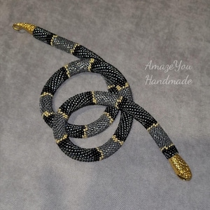 Puede incluir: Un collar con forma de serpiente de cuentas negras, grises y doradas. La serpiente está enrollada y tiene una cabeza y una cola doradas. El texto "Amaze You Handmade" es visible en el fondo.