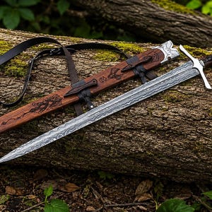 May include: A double-edged sword with a patterned Damascus steel blade, a silver crossguard, and a dark wood handle. The matching wooden scabbard features intricate carvings, silver metal accents, and an attached black leather carrying strap.