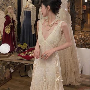 May include: A white lace dress with a plunging neckline and a fitted bodice. The dress has a flowing skirt with layers of lace and a delicate bow detail at the waist.