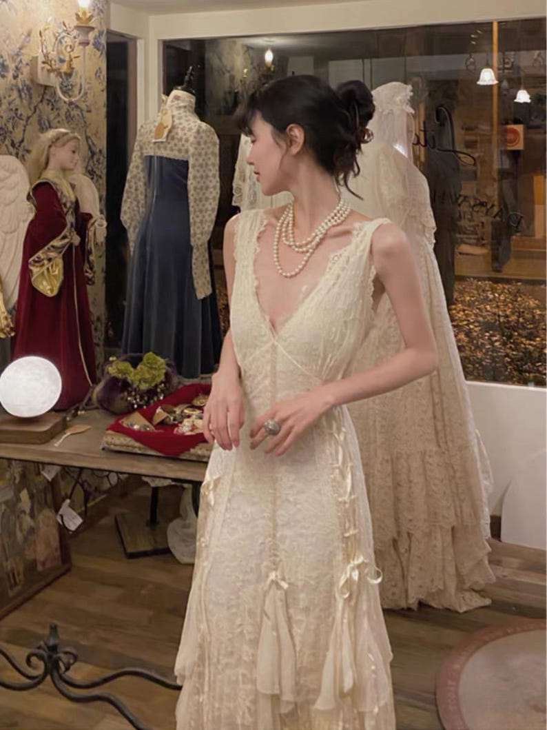 May include: A white lace dress with a plunging neckline and a fitted bodice. The dress has a flowing skirt with layers of lace and a delicate bow detail at the waist.