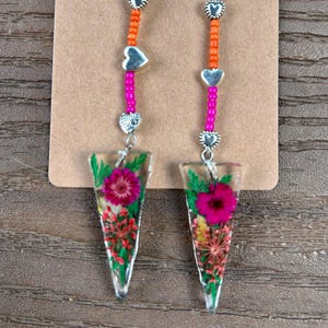 May include: A pair of dangle earrings with a floral design. The earrings feature clear, triangular pendants with pressed flowers. The beaded strands are pink and orange, with silver heart-shaped accents. The word "Glamorous" is printed at the top.