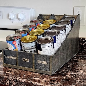 Può includere: Un organizer per lattine in legno con tre sezioni etichettate: Pomodori, Chili e Brodo. L'organizzatore è pieno di vari prodotti in scatola, con etichette visibili. La struttura in legno ha una finitura scura e invecchiata.