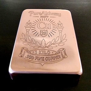 May include: A copper bar with the text "Pure Alchemy Mint" and "1 KILO .999 FINE COPPER" engraved on the surface. The bar features a design with torches, laurel leaves, and the chemical symbol "Cu" with the number 29.