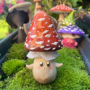 Peut inclure: Une figurine de champignon fantaisiste avec un chapeau rouge et des taches blanches, posée au milieu de la mousse verte. Le champignon a un visage souriant et des bras tendus. D'autres figurines de champignons colorés sont en arrière-plan.
