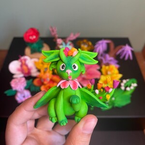 Peut inclure: Figurine de dragon vert faite à la main, avec une couronne de fleurs et des accents roses. Le dragon a de grandes oreilles et des ailes vertes, entouré de fleurs en argile colorées. La figurine est tenue dans une main.