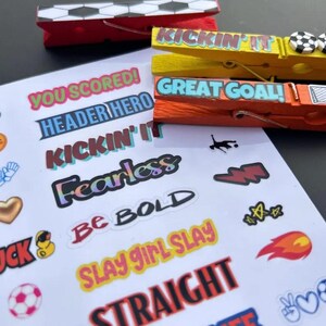 A white sheet of soccer-themed stickers with phrases such as You Scored, Header Hero, and Be Bold, alongside three decorated wooden clothespins in red, yellow, and orange. This set makes a thoughtful and unique gift for soccer players of all ages. These motivational clips can be attached to bags or jerseys to provide encouragement during a high-stakes tournament. They are a sweet way to show support for a favorite athlete.