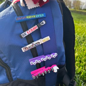 Cheer clothespin clips attached to a blue backpack, showing how finished cheer clips can decorate cheer bags and team backpacks. Features motivational phrases like “Cheer Loud,” “Be Loud,” “Fearless,” and custom bead clips, perfect for cheerleading team gifts, spirit pins, and competition favors.