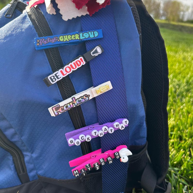 Cheer clothespin clips attached to a blue backpack, showing how finished cheer clips can decorate cheer bags and team backpacks. Features motivational phrases like “Cheer Loud,” “Be Loud,” “Fearless,” and custom bead clips, perfect for cheerleading team gifts, spirit pins, and competition favors.