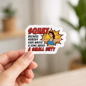 A hand holding a white sticker with a comic-book style illustration of a muscular woman flexing her biceps. This graphic sticker is part of the funny female weightlifting stickers collection, perfect for a power woman girl aesthetic.